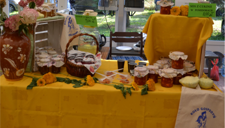 Na stole, przykrytym żółtym obrusem, leżą małe słoiczki z produktami w środku, stoi wazon z kwiatami, wiklinowy koszyk.