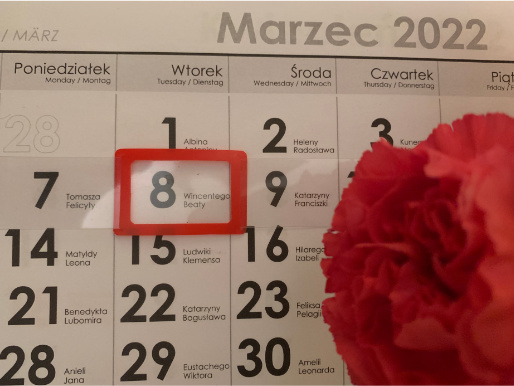 Kalendarz ścienny z miesiącem marzec. Zaznaczony dzień 8 marca. Obok leży czerwony kwiatek.