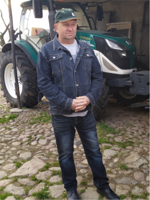 Mężczyzna w podeszłym wieku (Bronisław Zybała) stoi przed ciągnikiem rolniczym na brukowej kostce.