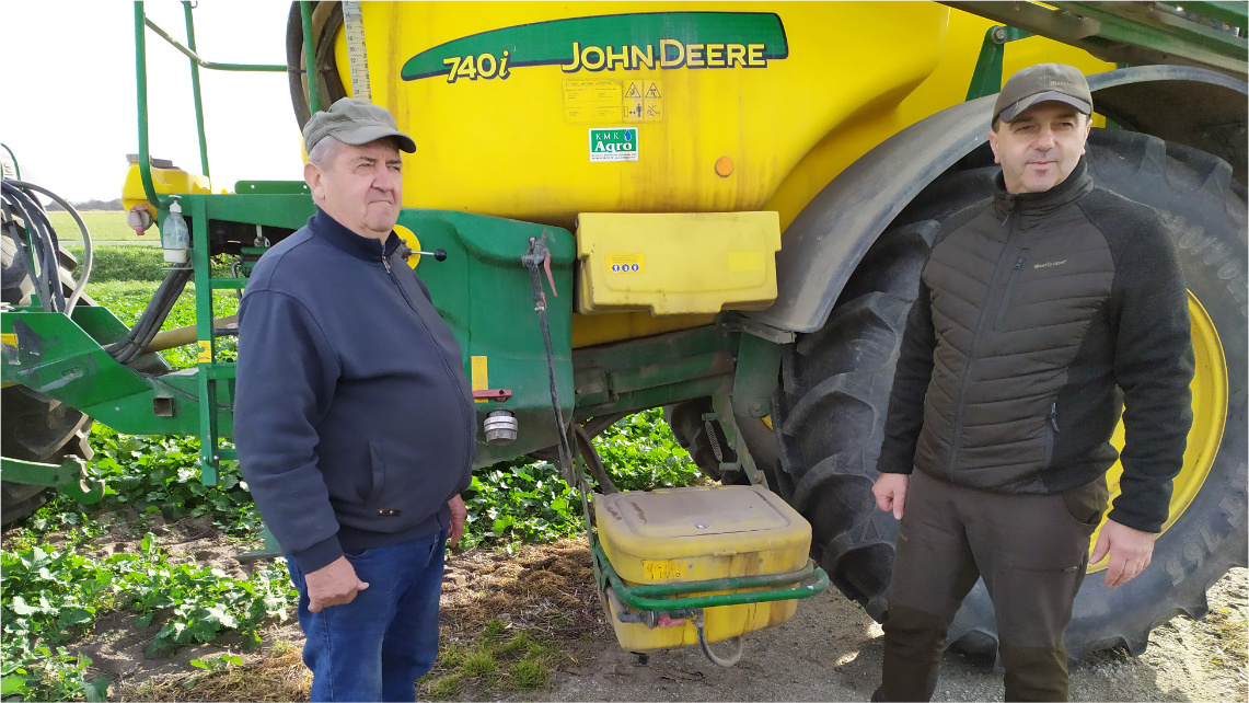 Piotr Łukaszewski i jego ojciec stoją na polu przed maszyną rolniczą.