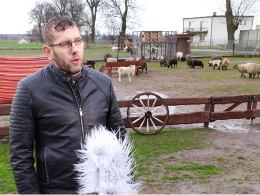 Pochmurny dzień. Młody mężczyzna - Przemysław Bilecki - wypowiada się dla ekipy TVP3 Poznań. W tle widać zwierzęta.