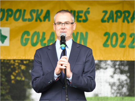 Mężczyzna w średnim wieku, Maciej Szłykowicz, stoi na scenie i przemawia przez mikrofon.