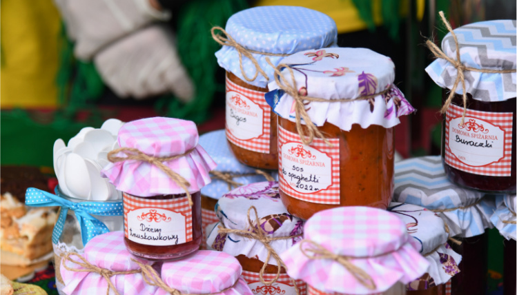 Konferencja: „Identyfikacja i promocja żywności wysokiej jakości szansą dla lokalnych producentów” Stos słoików z domowymi przetworami, takimi jak bigos czy sos do spaghetti. Słoiki mają kolorowe papierowe nakładki na wieka.