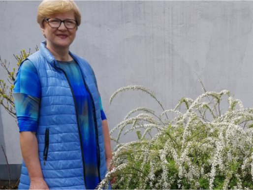 Po lewej stronie stoi pani Ewa ubrana w niebieską bluzkę i ocieplaną kamizelkę. Pozuje do zdjęcia. Obok niej znajduje się bujny krzew z białymi kwiatami.