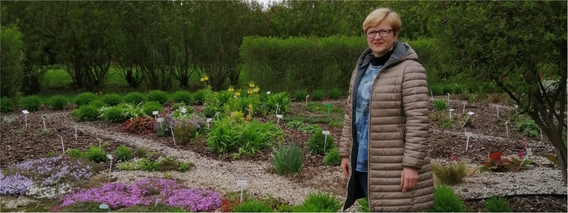 Na pierwszym planie pozuje Ewa Tuliszka. W tle znajduje się ogródek z kwiatami w różnych kolorach.
