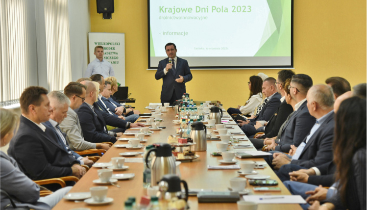 Organizatorem przyszłorocznych Krajowych Dni Pola jest Wielkopolski Ośrodek Doradztwa Rolniczego w Poznaniu Sala konferencyjna. Przy długim stole nakrytym zastawą do kawy siedzą ludzie po obu stronach. Na szczycie stołu stoi mężczyzna w garniturze i przemawia. Za nim jest ekran z wyświetloną prezentacją na temat Krajowych Dni Pola 2023.