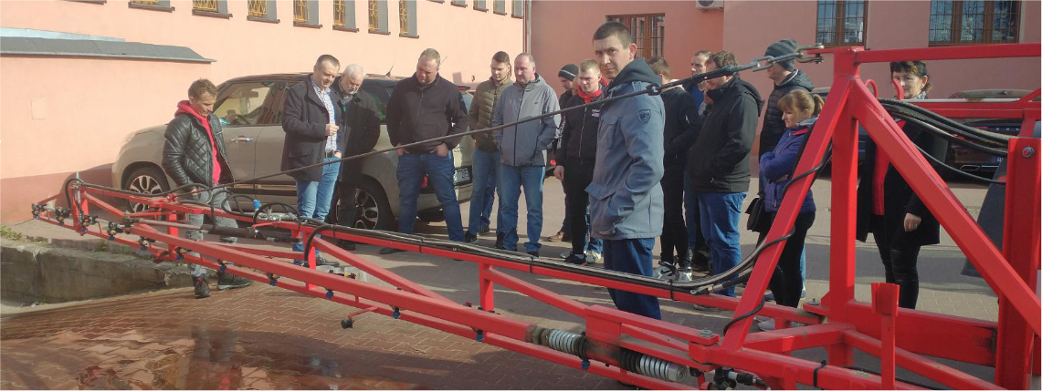 Przed budynkiem stoi grupa ludzi, obok nich jest maszyna do pryskania upraw.