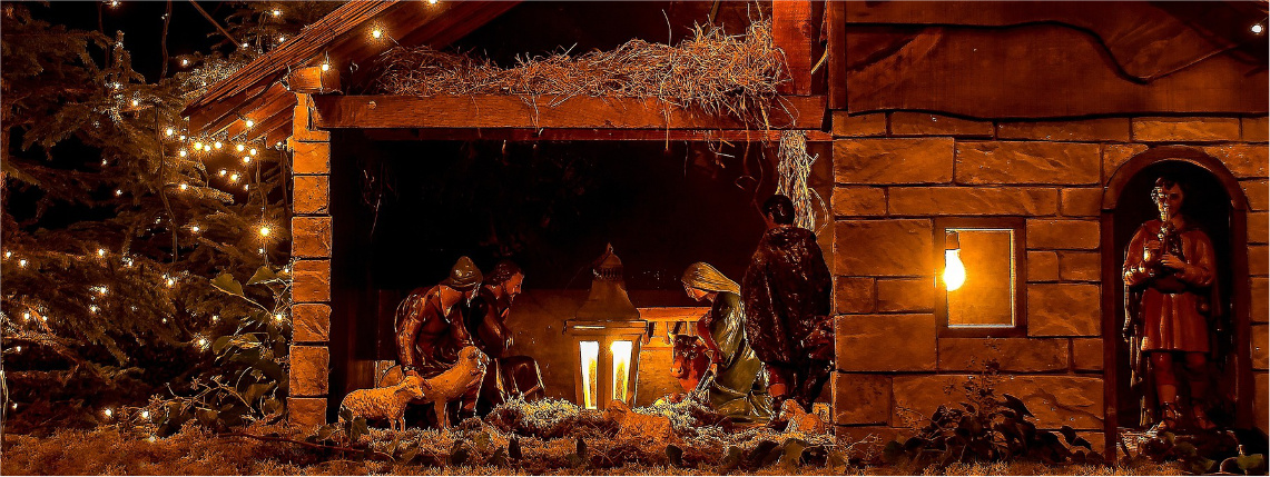 Zdjęcie przedstawiające szopkę bożonarodzeniową z figurkami świętej rodziny. Obok jest drzewko z lampkami. Zdjęcie jest przyciemnione i nastrojowe.