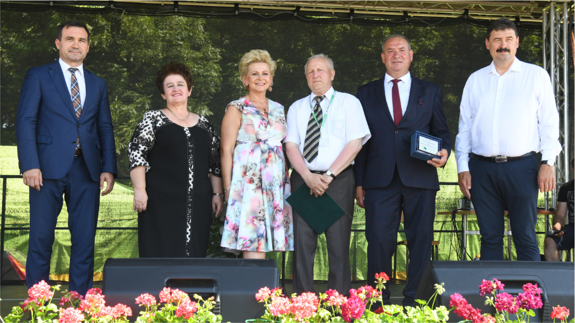 Na scenie w rzędzie stoi sześć osób, elegancko ubranych, zwróconych przodem. Z lewej jest mężczyzna, dalej dwie kobiety i trzech mężczyzn. Przed nimi stoją różowe kwiaty rabatowe.