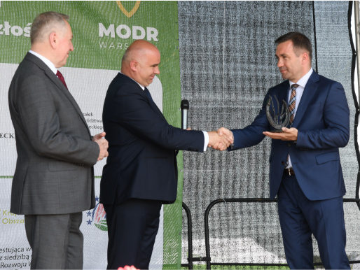 Przekazanie symbolicznej podkowy na ręce dyrektora WODR, Jacka Sommerfelda Na scenie stoi trzech mężczyzn w garniturach. Dwóch z nich wymienia uścisk dłoni i patrzy na siebie.