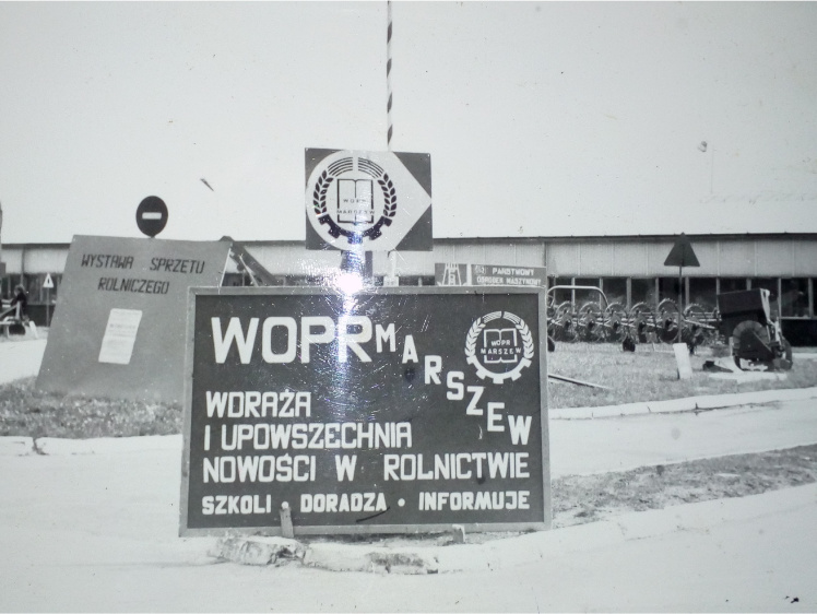 Czarno-białe zdjęcie. Tablica z napisem "WOPR Marszew" stoi na terenie targów. W tle widać budynek