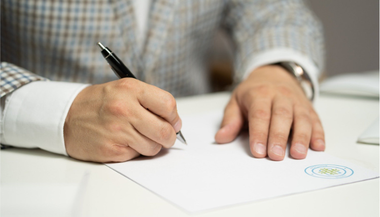 Zbliżenie na ręce mężczyzny ubranego w garnitur, siedzącego przy stole. Trzyma w dłoni długopis i pisze na kartce.