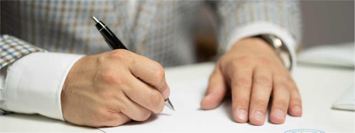 Zbliżenie na dłonie leżące na stole na kartce papieru. Jedna dłoń trzyma długopis.