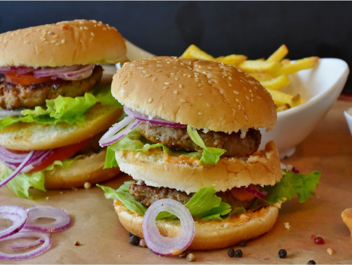 Dwa piętrowe hamburgery, przełożone są mięsem, cebulą i sałatą. W tle widać frytki. Przed hamburgerem leży pokrojona w plasterki czerwona cebula.