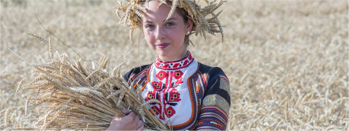 Młoda kobieta w ludowym stroju stoi na polu między uprawami. W ręku trzyma kłosy zboża.