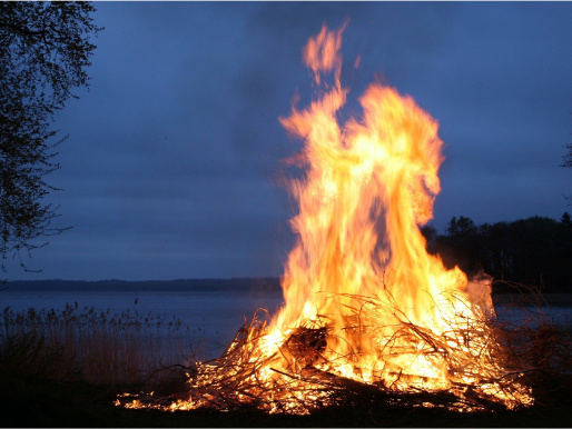 Wieczór, z bliska widać rozpalone ognisko. Ogień wznieca się na kilka metrów. W tle znajduje się jezioro.