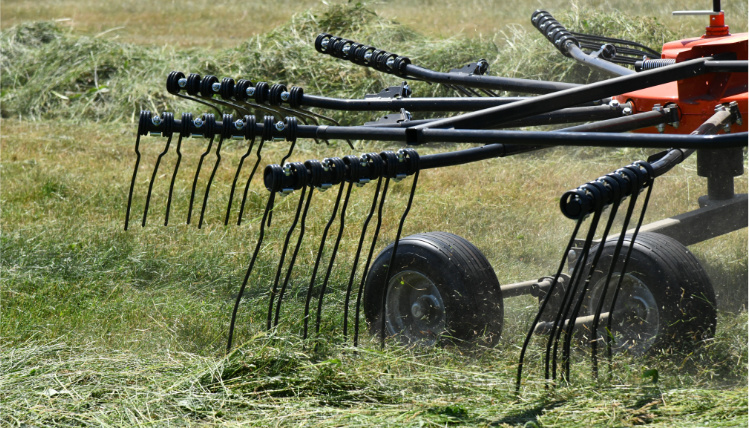 Maszyna rolnicza pracuje na polu. Zbliżenie na koła oraz podłużne elementy do zbierania trawy.