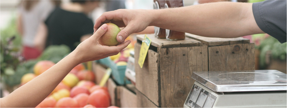 Stoisko handlowe z owocami. Nad koszami jabłek jedna osoba podaje drugiej jabłko. Widać jedynie ręce tych osób.