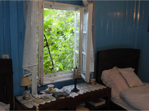 Wnętrze pomieszczenia w starym domu. Ściany są niebieskie, znajdują się tu stare meble. Za otwartym oknem widać zielone drzewa.