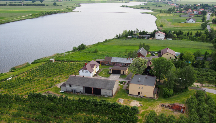Malowniczy krajobraz gospodarstwa agroturystycznego "Owocowy Raj Nad Zalewem" Widok z lotu ptaka na gospodarstwo położone między zielonymi polami. Obok widać duży zalew.