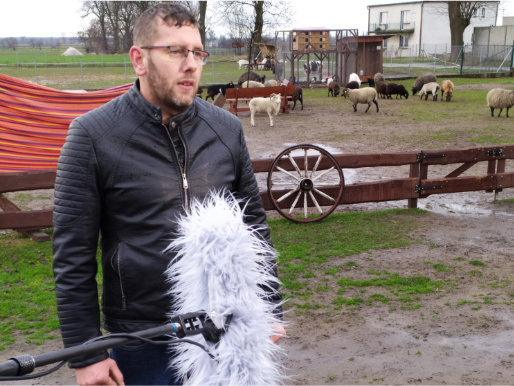 Pochmurny dzień. Młody mężczyzna, Przemysław Bilecki, stoi i wypowiada się do mikrofonu, który jest przed nim. W tle widać zwierzęta.