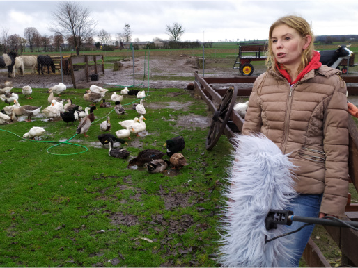 Pochmurny dzień. Młoda kobieta, Monika Bilecka, stoi na trawie i wypowiada się do mikrofonu, który jest przed nią. W tle widać zwierzęta na trawie.