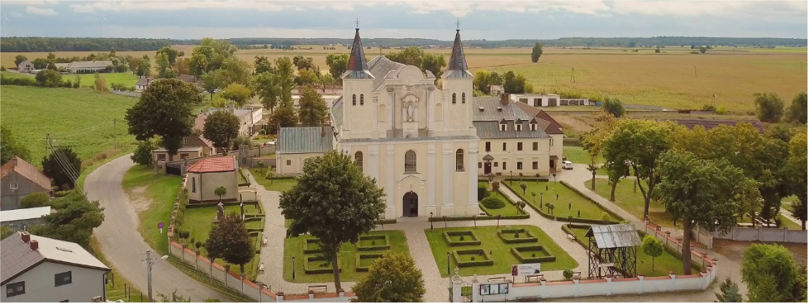 Widok z góry na sanktuarium - biały budynek z dwiema wieżami. Wokół niego jest zielony teren ogrodzony płotem. W tle widać domy, drzewa i las.