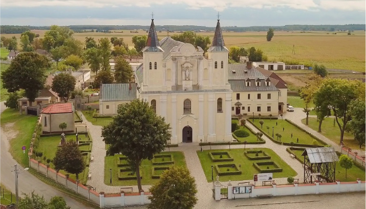 Widok z góry na sanktuarium - biały budynek z dwiema wieżami. Wokół niego jest zielony teren ogrodzony płotem. W tle widać domy, drzewa i las.