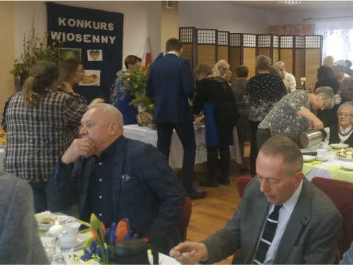 W degustacji potraw uczestniczyło wiele osób Wnętrze sali. Na pierwszym planie widać dwóch mężczyzn w średnim wieku siedzących przy stołach do degustacji potraw. W tle widoczni są inni ludzie, którzy stoją przy stole.