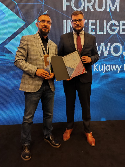 Maciej Zacharczuk (po lewej) i Adam Fojud (po prawej), informatycy WODR, stoją z nagrodą i dyplomem na scenie. Za nimi jest niebieskie tło.