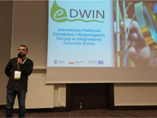 Maciej Zacharczuk stoi i wygłasza wykład. W tle telebim, na którym widać logo projektu eDWIN.
