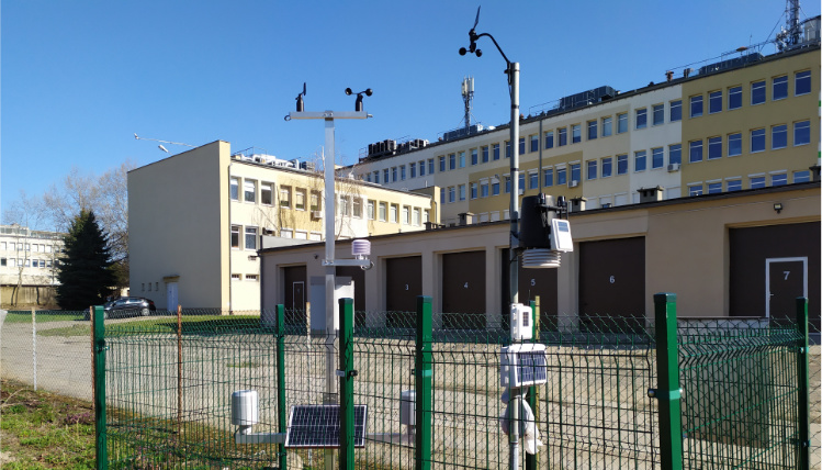 Słoneczny dzień. Przed budynkiem WODR w Poznaniu stoi stacja meteorologiczna. Jest ona ogrodzona płotem.