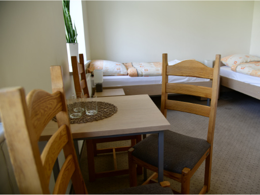 Wnętrze pokoju. Dwa krzesła stoją przy kwadratowych stole. W tle widać dwa łóżka pojedyncze. 