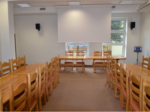 Wnętrze sali konferencyjnej. Drewniane stoły i krzesła stoją w dwóch rzędach. W tle widać opuszczoną tablicę, na której można wyświetlać prezentacje.