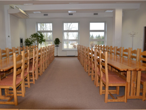 Wnętrze pomieszczenia. Drewniane stoły i krzesła stoją w dwóch rzędach. W tle widoczne są okna.