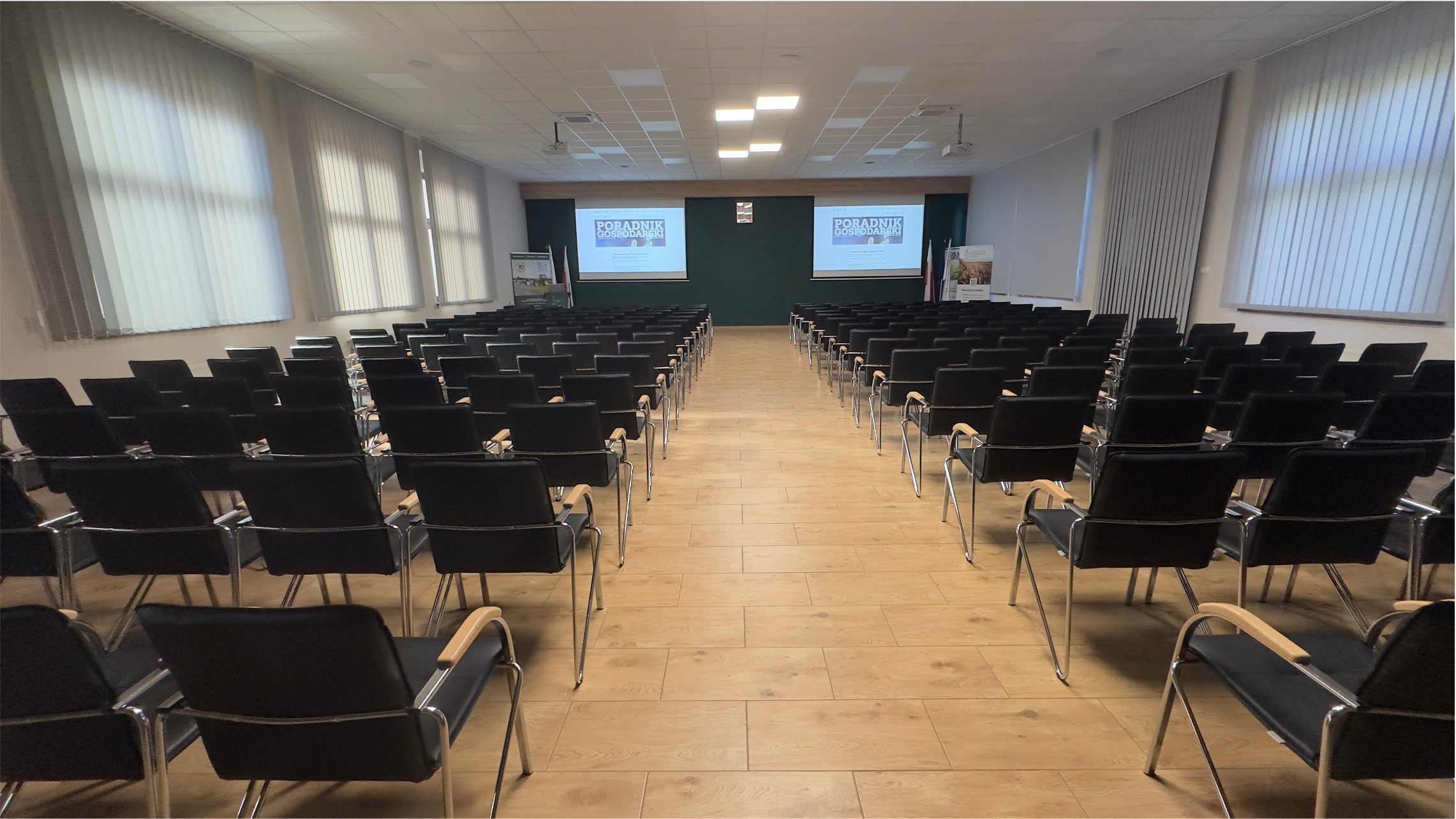 Wnętrze sali konferencyjnej w Sielinku Wnętrze sali konferencyjnej. Drewniane stoły i krzesła stoją w dwóch rzędach. W tle widać okna.