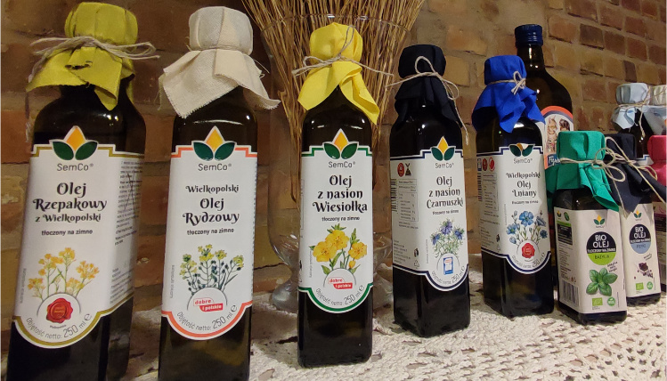 Szklane butelki z olejem stoją na obrusie na półce przed ceglaną szklaną. Na butelkach olejów są etykiety z informacją o rodzajach olei.