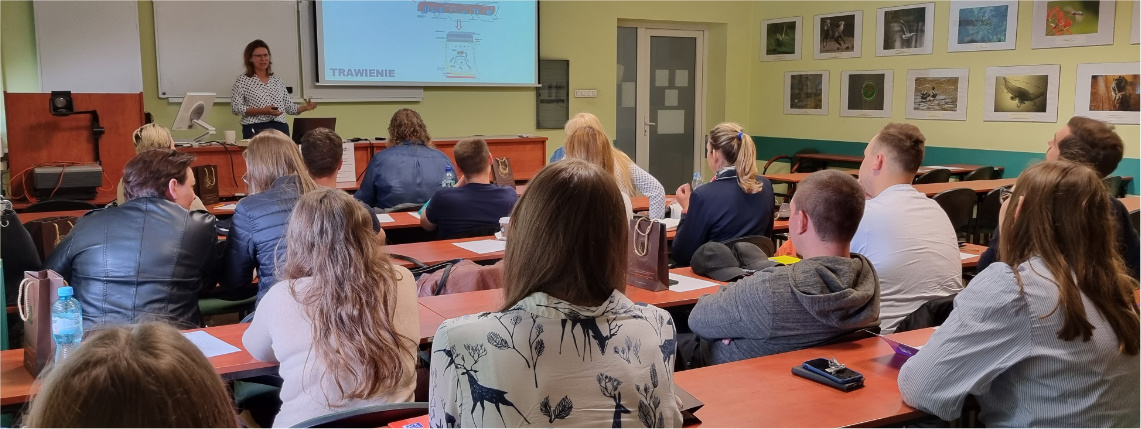 Pierwsze szkolenie z cyklu "Akademia żywienia krów mlecznych - wyzwania współczesnego producenta mleka" Sala wykładowa. Przy biurkach siedzą słuchacze, którzy są widoczni od tyłu. Przed nimi stoi wykładowca i omawia prezentację, która jest wyświetlona na ekranie.