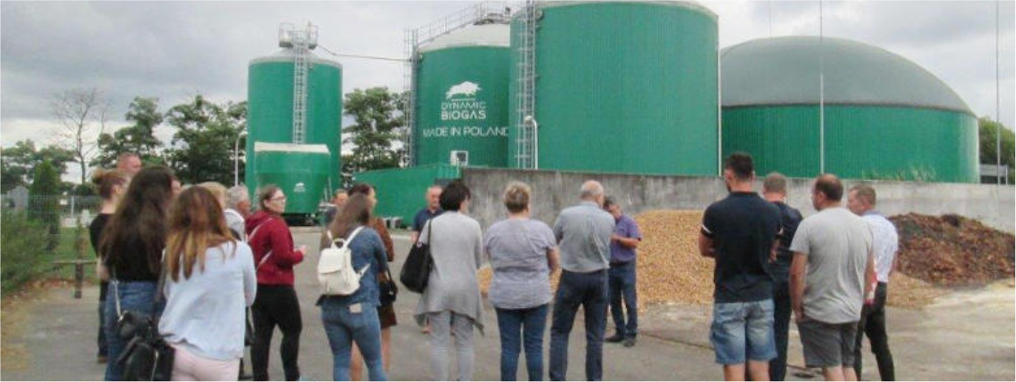 Pochmurny dzień. Grupa kilkunastu osób, widocznych od tyłu, stoi kilkadziesiąt metrów przed biogazownią w Przybrodzie.