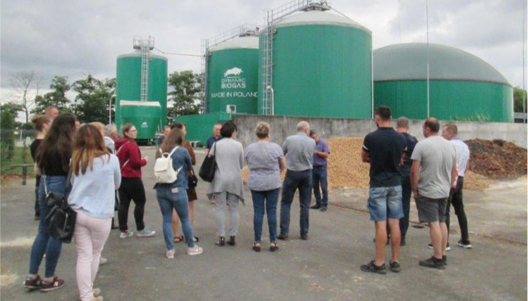 Wyjazd do biogazowni w Przybrodzie został zorganizowany w ramach Sieci na rzecz innowacji w rolnictwie i na obszarach wiejskich Pochmurny dzień. Grupa kilkunastu osób, widocznych od tyłu, stoi kilkadziesiąt metrów przed biogazownią w Przybrodzie.