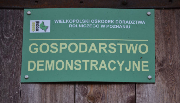 Na drewnianej ścianie wisi zielona tabliczka z żółtym napisem "Gospodarstwo demonstracyjne" oraz logiem WODR.