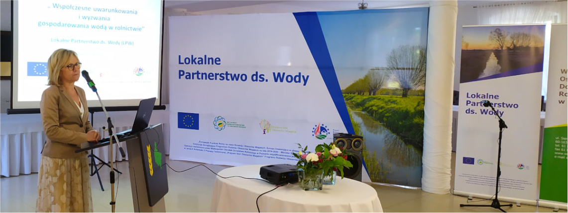 Wnętrze pomieszczenia. Kobieta w średnim wieku - Wiesława Nowak - stoi przed mównicą z logo WODR. W tle widać prezentację na telebimie i napis "Lokalne Partnerstwa ds. Wody".