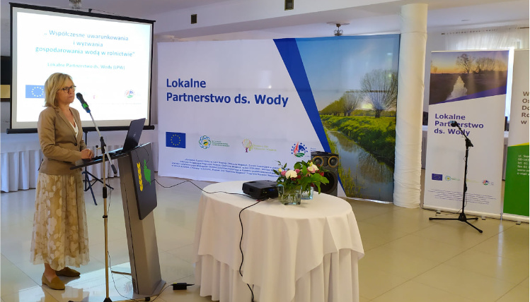 Wnętrze pomieszczenia. Kobieta w średnim wieku - Wiesława Nowak - stoi przed mównicą z logo WODR. W tle widać prezentację na telebimie i napis "Lokalne Partnerstwa ds. Wody".