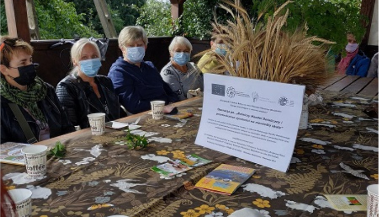 Na zdjęciu widoczne są kobiety siedzące przy stole w gospodarstwie Aleksandry i Mieczysława Babalskich. Na środku stoi biała kartka z informacją o realizacji operacji.