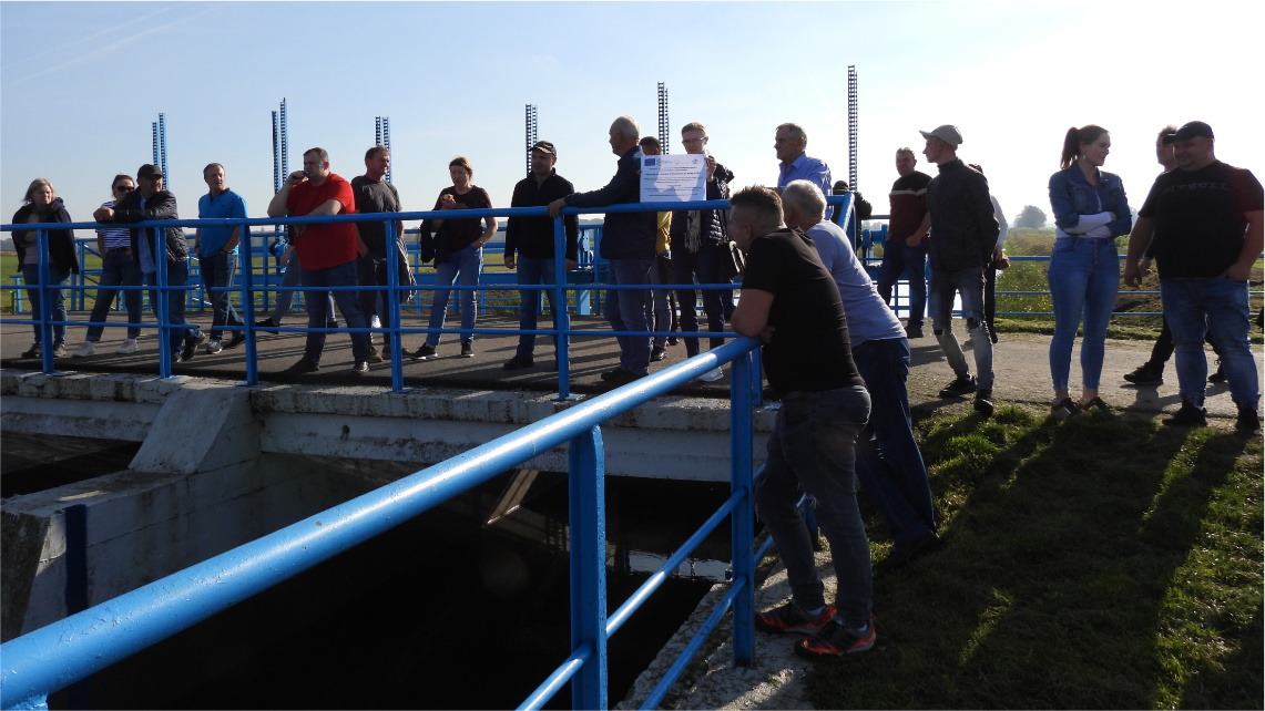 Słoneczny dzień. Grupa ludzi stoi przy zbiorniku wodnym i opiera się o niebieską barierkę.