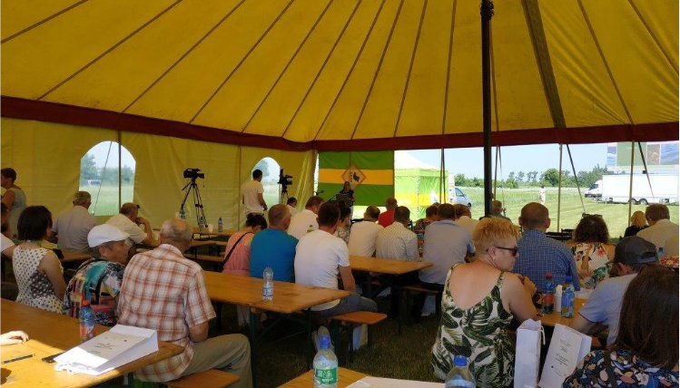 Wnętrze dużego namiotu. Przy stołach, na ławkach, siedzi kilkadziesiąt osób, które słuchają wykładu. W tle widoczna jest wykładowczyni.