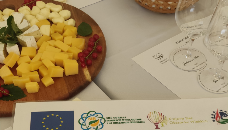 W dniach 28-30 czerwca 2022 roku zorganizowany został wyjazd studyjny dla grupy 23 osób Na stole stoi drewniana taca z kawałkami owoców. Obok niej stoją kieliszki do wina, a przed nią leży biała kartka z czterema różnymi logotypami.