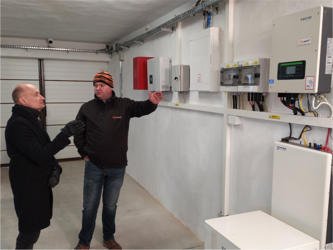 Dwaj mężczyźni w średnim wieku, Janusz Michałowicz i Marcin Krawczyk, stoją wewnątrz pomieszczenia przed urządzeniami energetycznymi, które wiszą na ścianie.