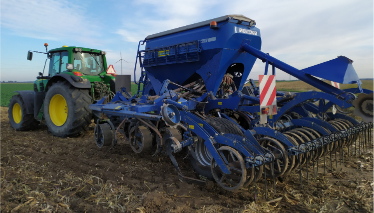 Pochmurny dzień. Zielony ciągnik rolniczy z przyczepioną do niego wielką niebieską maszyną do prac rolniczych stoi na polu.