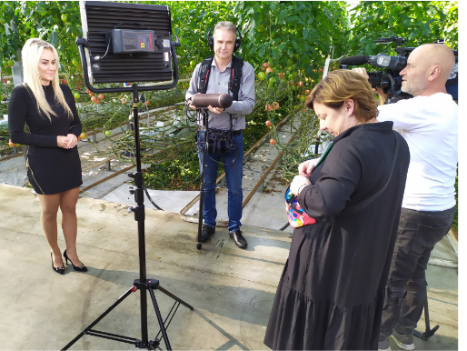 Wnętrze szklarni. Młoda kobieta, Agnieszka Kaźmierczak, stoi z ekipą telewizyjną obok pnączy, na których rosną pomidory. Przeprowadzany jest wywiad.